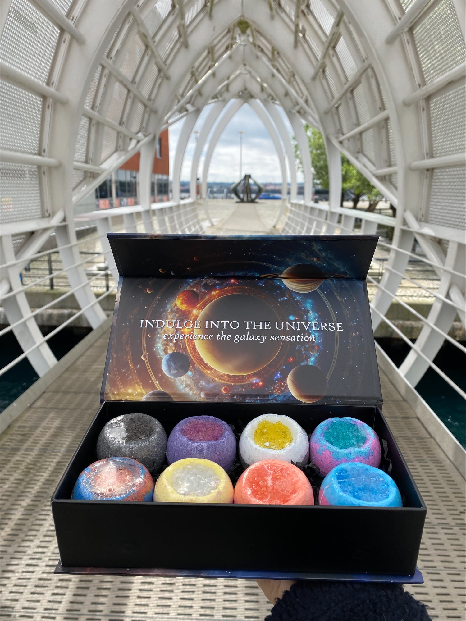 Box of colourful galaxy-themed bath bombs displayed on a modern bridge with cosmic packaging that reads “Indulge into the universe”