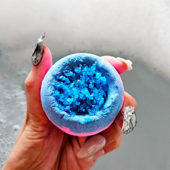Supernova rosemary bath bomb with vibrant blue crystals held over a bubble bath